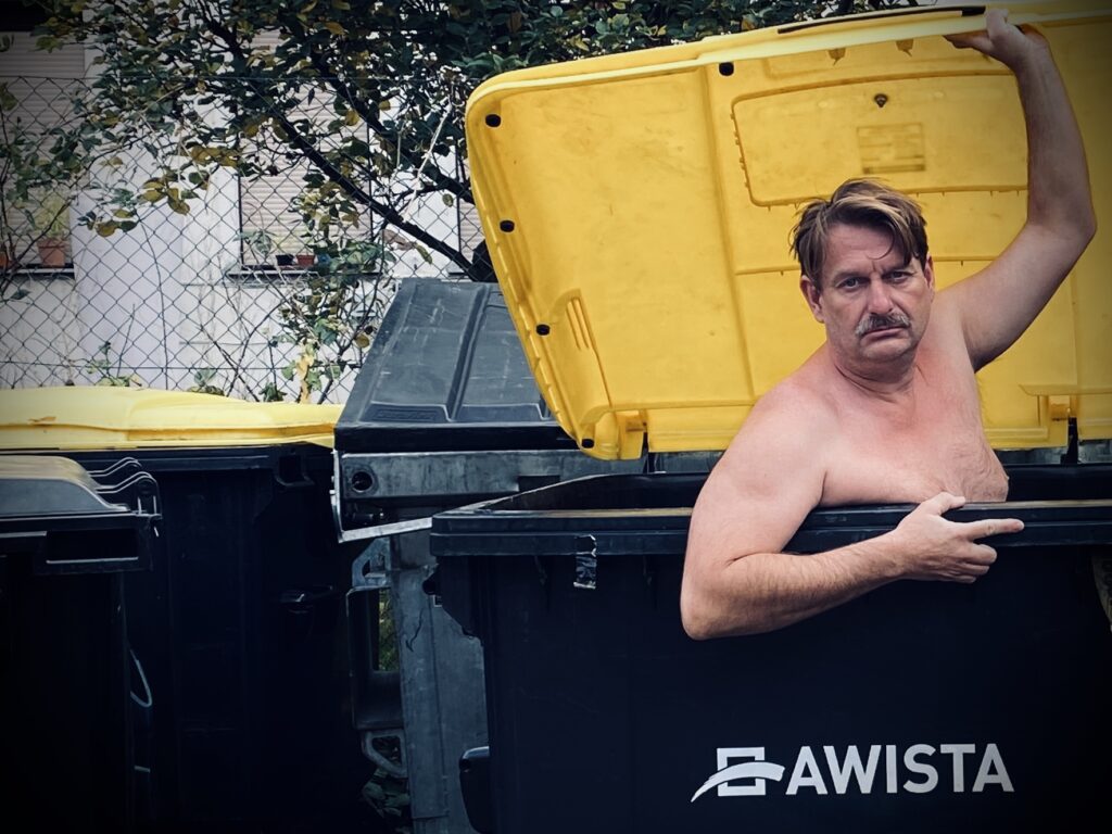 Peter Trabner sitzt nackt in einer Mülltonne und hebt den Deckel an